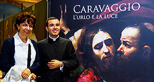 Don Elenio Abis e la Professoressa Alessandra pasolina in posa con nello sfondo il quadro di Caravaggio, locandina della mostra "L'Urlo e la Luce"